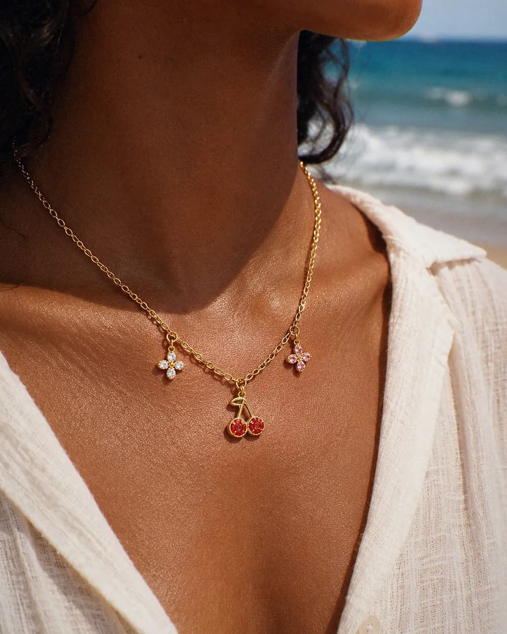 Collier à charms dorés — cerises rouges et deux fleurs en cristal — porté face à la plage